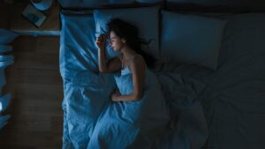Top View of Young Woman Sleeping Cozily on a Bed in Bedroom at Night. Blue Nightly Colors with Cold Weak Lamppost Light Shining Through the Window.