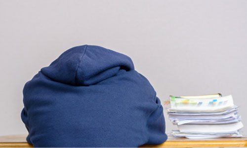 View from behind of person in hoodie with head on desk with pile of papers next to them